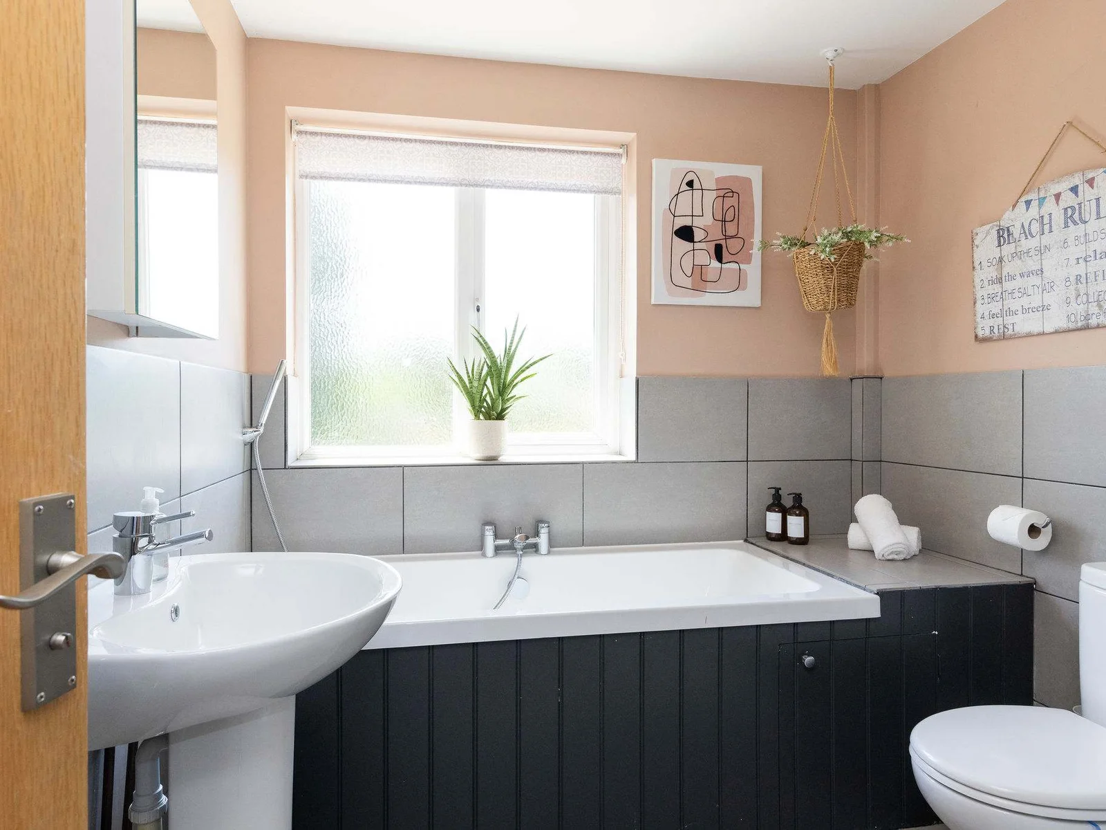 A fresh, well-appointed family bathroom with a full-size bath, sleek dark cabinetry, soft blush walls and contemporary grey tiling, all brought together with plenty of natural light.