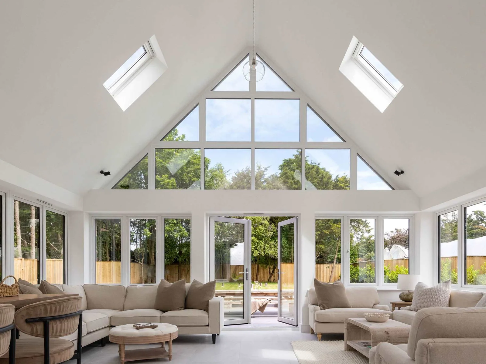 Sun-drenched, open-plan living space featuring a vaulted ceiling with skylights, a striking triangular gable window framing a lush green garden view, plush cream sectionals, and direct patio access.