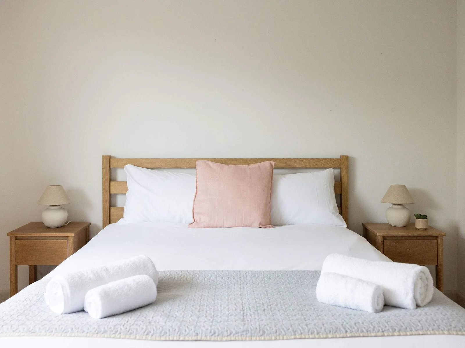 Bright, calming bedroom featuring a comfortable King bed, natural wood bedside tables, soft neutral bedding with a blush accent cushion, matching bedside lamps and fresh towels ready for your stay.