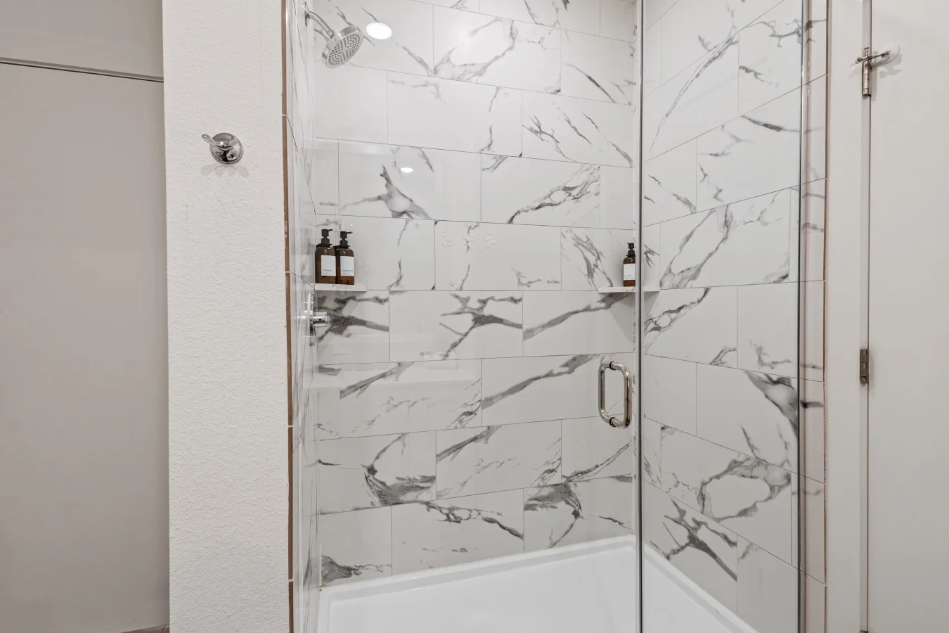 Refreshing modern shower featuring sleek glass door, elegant marble-patterned tiles, and convenient wall shelves for your essentials.