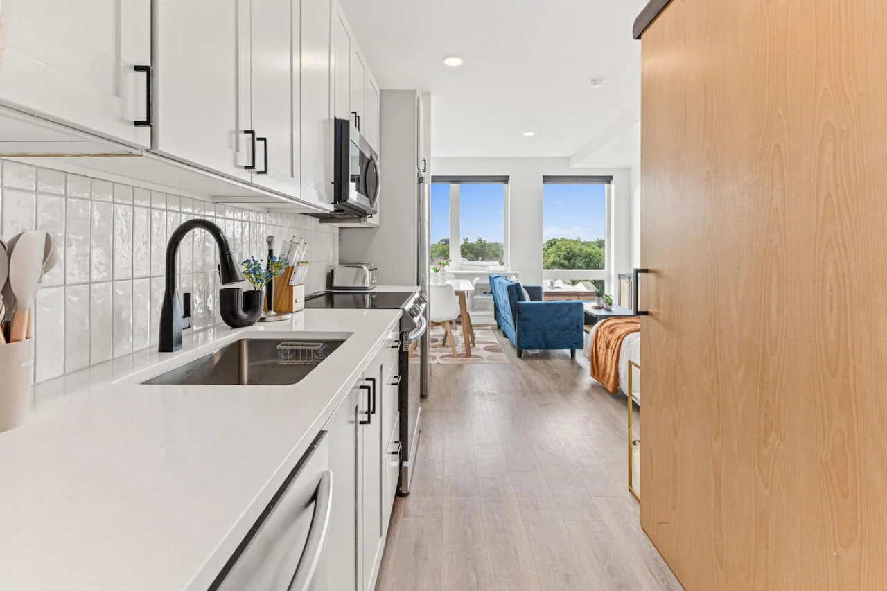 This modern kitchen features a spacious island with cabinetry, sleek white countertops, stainless steel appliances, open shelving, and stylish lighting.