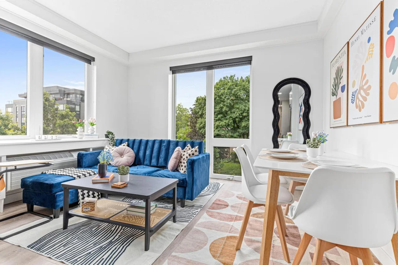 Cozy living room w/ couch & Throw pillows - Coffee table - Area rug - Wall mounted mirror - Large windows allow for Natural light - Room darkening shades - Dining table w/ 4 chairs a place settings - Side table - Books.