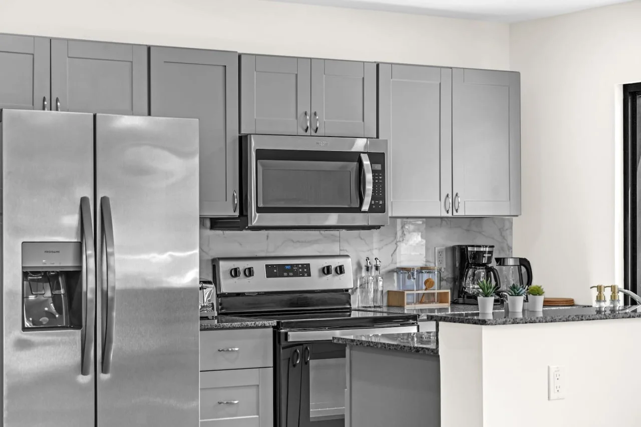 This modern kitchen features a spacious island with cabinetry, sleek white countertops, stainless steel appliances, open shelving, and stylish lighting.