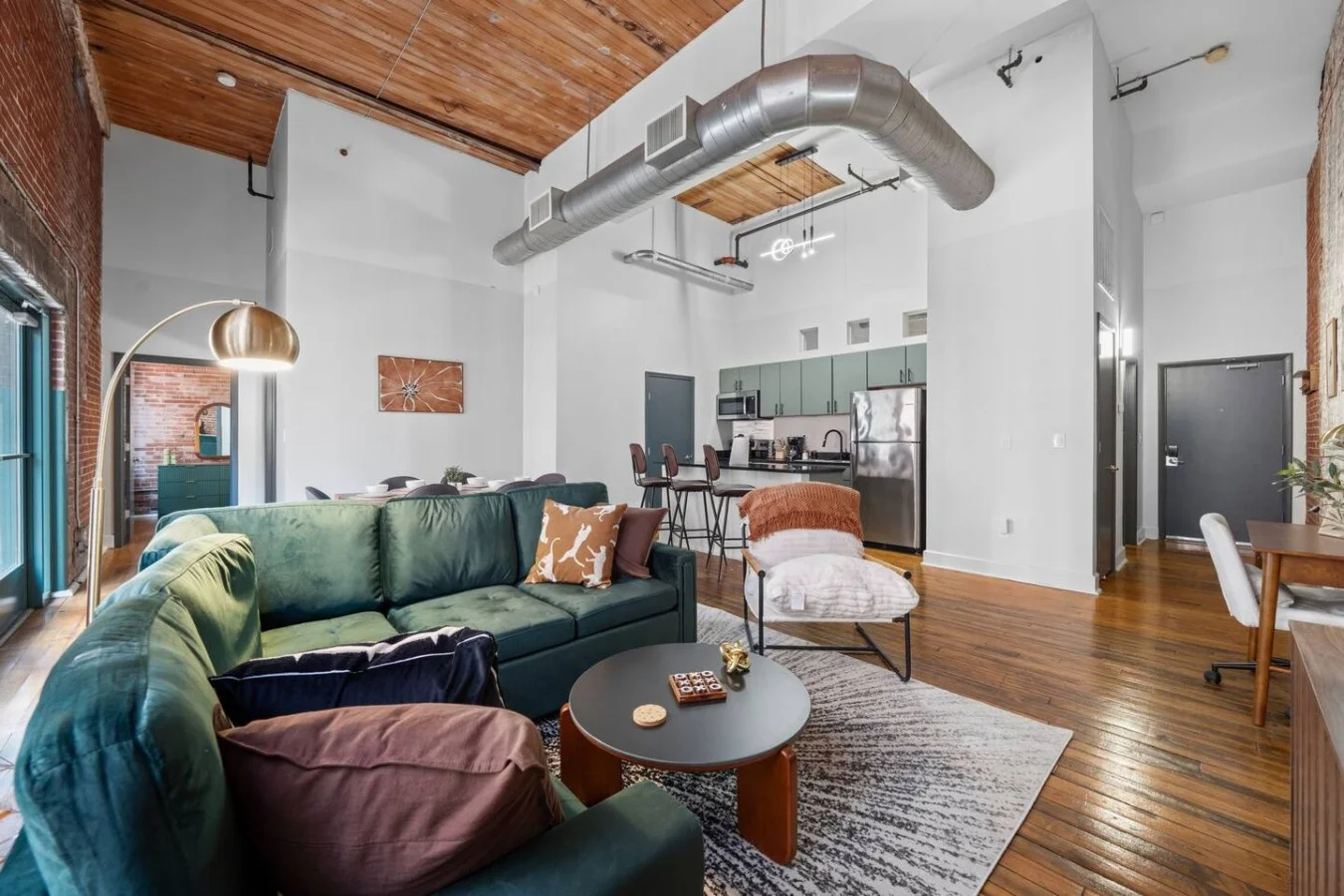 Bright, modern loft with high ceilings, exposed brick, full kitchen, dining area, workspace, and cozy lounge. Warm wood and big windows create an inviting, open feel.