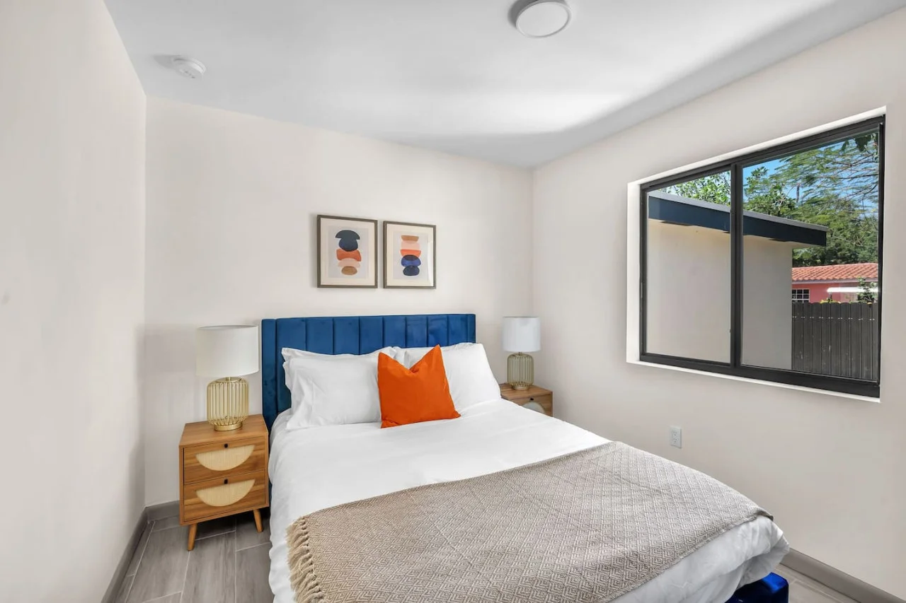 Bright bedroom featuring a soft, cushioned blue headboard, vibrant orange accent pillows, and natural lighting through a large window.