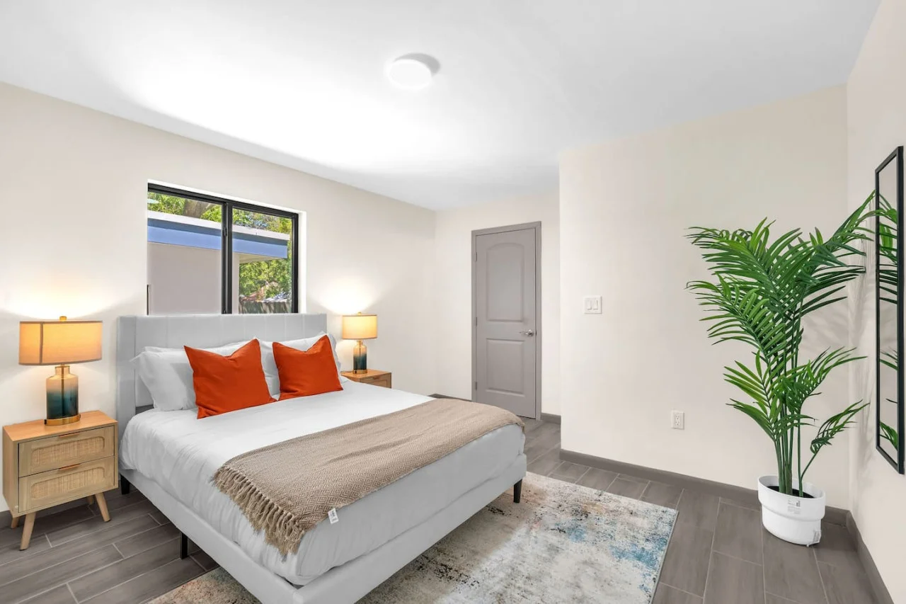 A bright and inviting bedroom with a modern design, featuring crisp white bedding, vibrant orange accent pillows, and natural light streaming through the large windows.