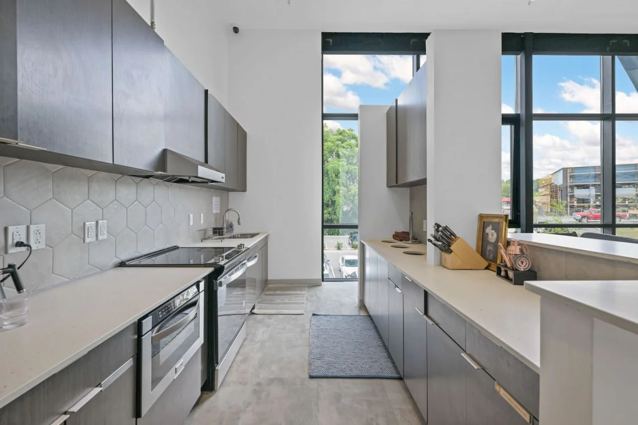 This modern kitchen features a spacious island with cabinetry, sleek white countertops, stainless steel appliances, open shelving, and stylish lighting.