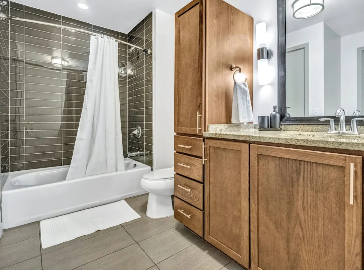 This bright and welcoming bathroom invites you to freshen up in comfort. Enjoy the forest-inspired shower curtain that adds a calming touch to your routine, complemented by the cozy rug underfoot and sparkling clean fixtures.