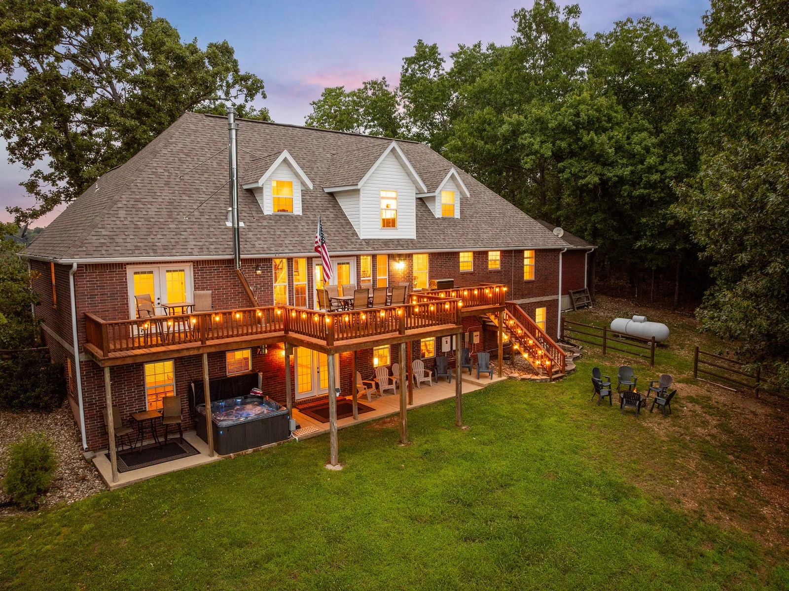 At dusk the whole property lights up: string lights on the upper deck, warm glow from the lower patio, fire already crackling in the pit. Three levels of evening atmosphere.