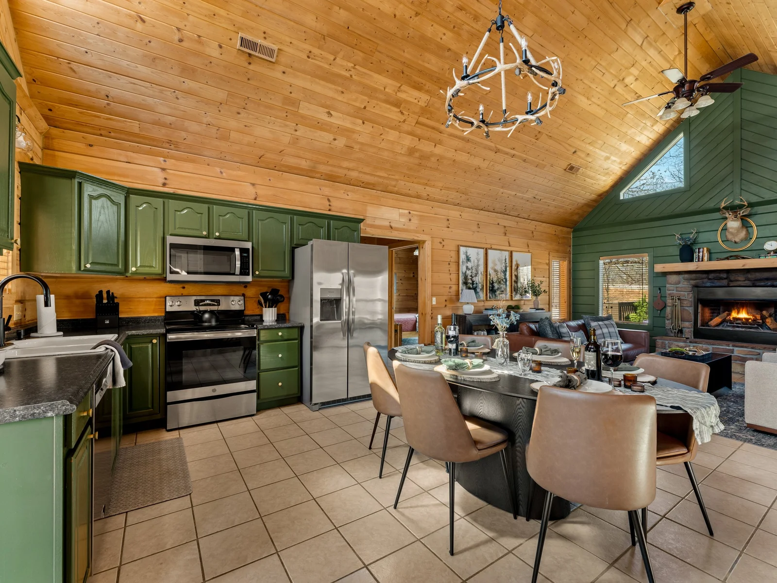 Kitchen with stainless appliances beside the dining table. An antler chandelier hangs over dinner while the fire glows across the room.