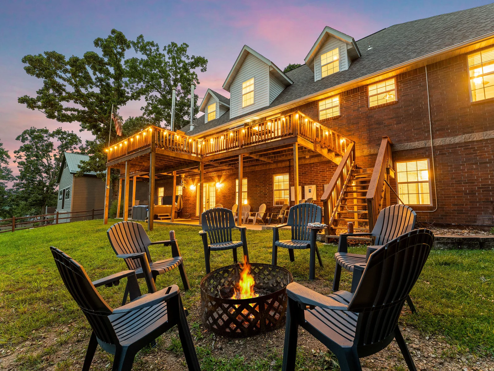 The full rear of the house at dusk — string lights wrapping the deck, the fire pit glowing below, the lake visible through the trees. This is the whole property in one shot.