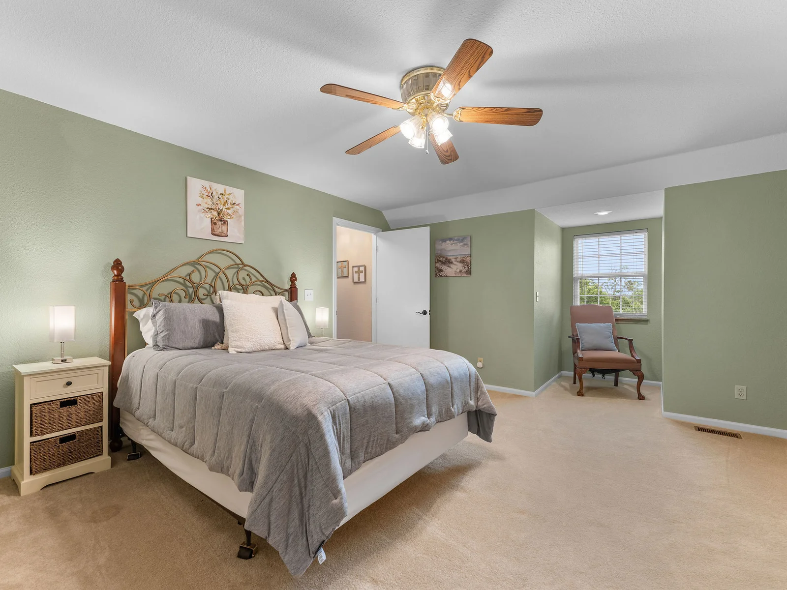 The upstairs guest bedroom: a queen bed, sage green walls, carpeted floor, and a sitting nook with natural light. Quiet and comfortable at the top of the house.