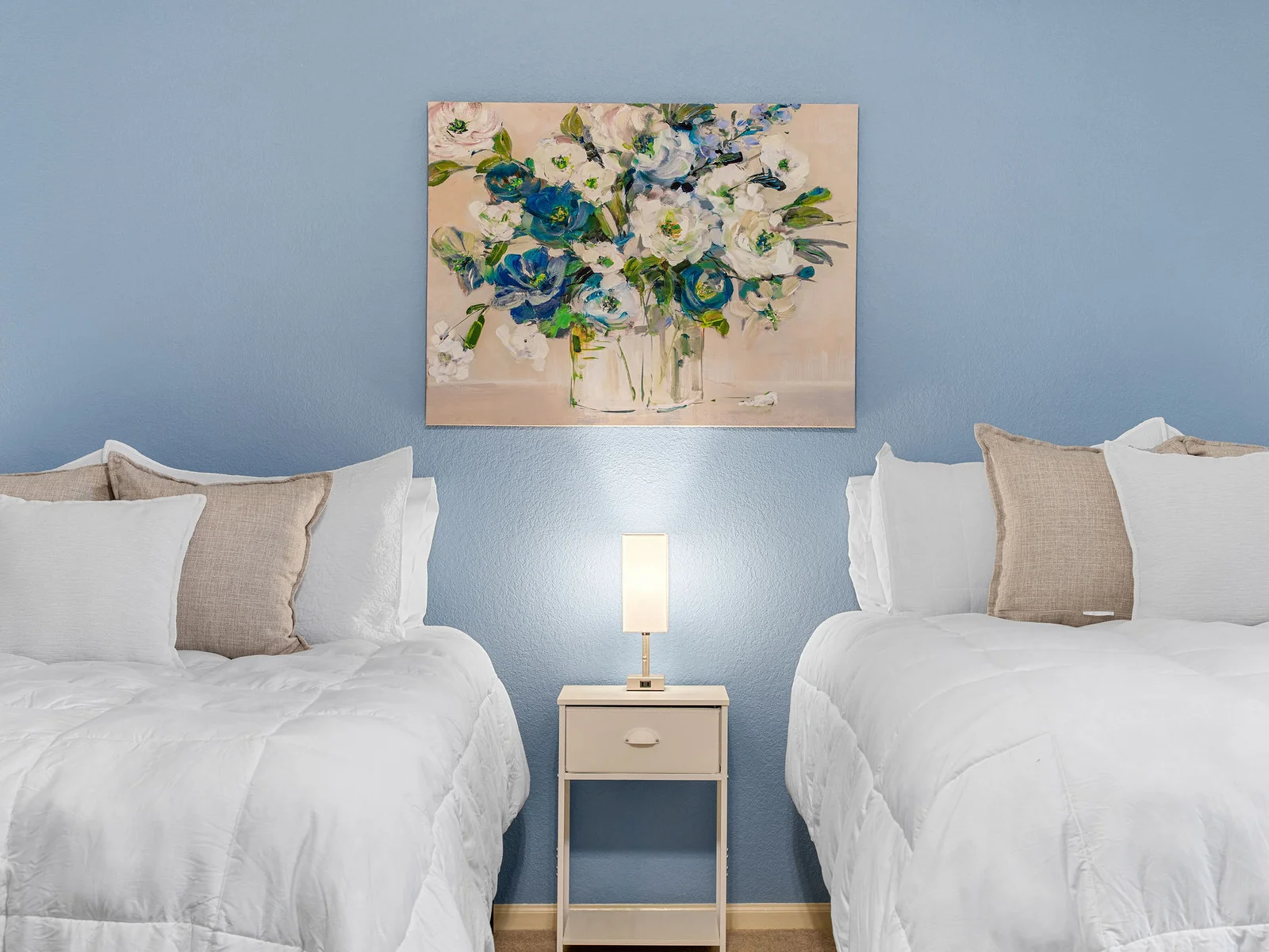 The twin-bed room on the upper floor sleeps two in full beds with a blue accent wall, framed floral art, and a small reading corner. Great for kids or friends sharing.
