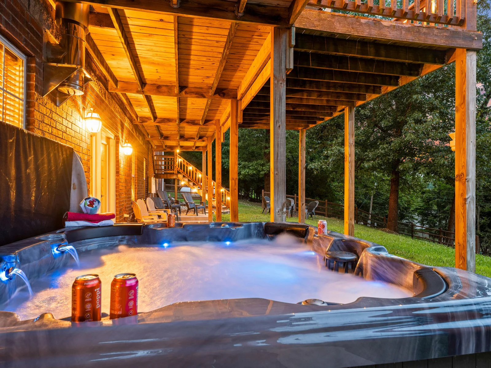 Amber light from the deck above, steam rising from the tub, the lake through the trees in the background — the private hot tub from the lower patio at dusk.